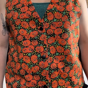 May include: A black sleeveless vest with a pattern of orange jack-o'-lantern faces on a black background. The vest has a V-neck and three black buttons.