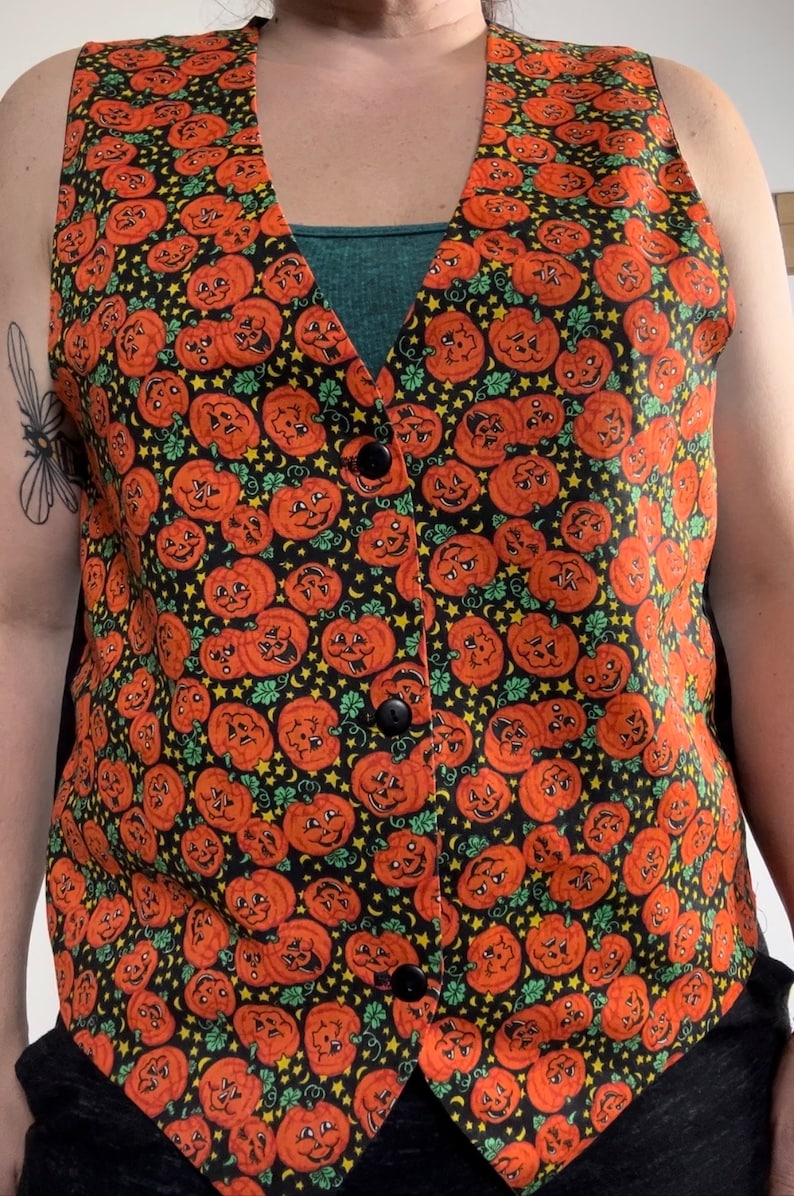 May include: A black sleeveless vest with a pattern of orange jack-o'-lantern faces on a black background. The vest has a V-neck and three black buttons.
