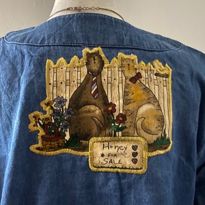 May include: Blue denim shirt with a patch of two cats wearing ties and a sign that says "Honey For Sale".