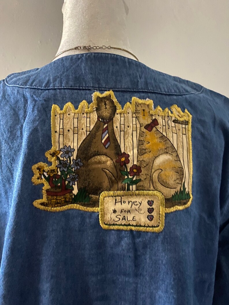 May include: Blue denim shirt with a patch of two cats wearing ties and a sign that says "Honey For Sale".