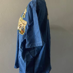 May include: A blue denim shirt with a colorful graphic print of a garden scene. The print includes a basket of flowers, a watering can, and a sign that says "Plant Sale".