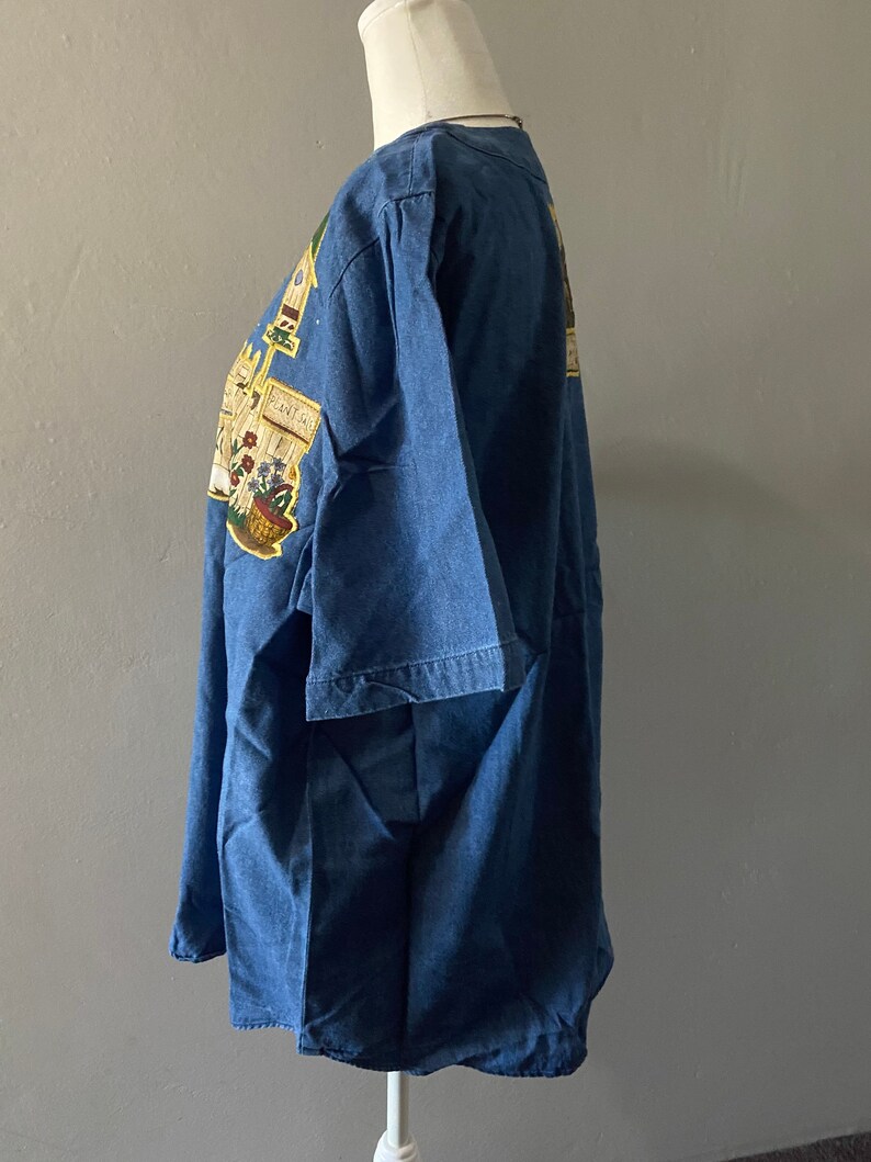 May include: A blue denim shirt with a colorful graphic print of a garden scene. The print includes a basket of flowers, a watering can, and a sign that says "Plant Sale".