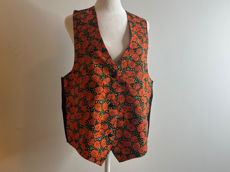 May include: A black vest with a pumpkin print in orange and green. The vest has a V-neck and a button closure.