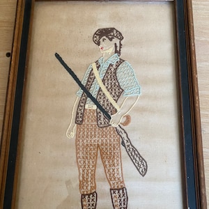 May include: A framed cross-stitch embroidery of a person wearing a brown hat and brown pants holding a rifle. The embroidery is done in shades of brown, blue, and white.
