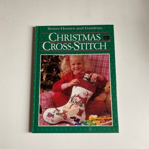May include: A book titled "Better Homes and Gardens Christmas Cross-Stitch" with a green cover and a picture of a young girl holding a Christmas stocking. The stocking is white with a colorful cross-stitch design of Santa Claus and other Christmas characters.
