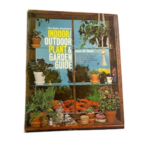 May include: A wooden display case with glass shelves, filled with potted plants and small decorative items. The case features a book titled "Indoor/Outdoor Plant & Garden Guide". The background shows a window with orange flowers.