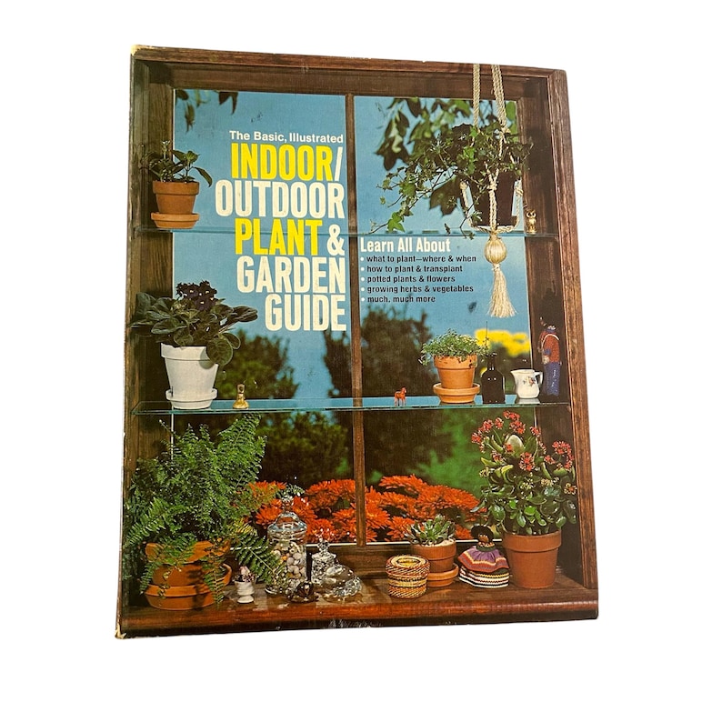 May include: A wooden display case with glass shelves, filled with potted plants and small decorative items. The case features a book titled "Indoor/Outdoor Plant & Garden Guide". The background shows a window with orange flowers.