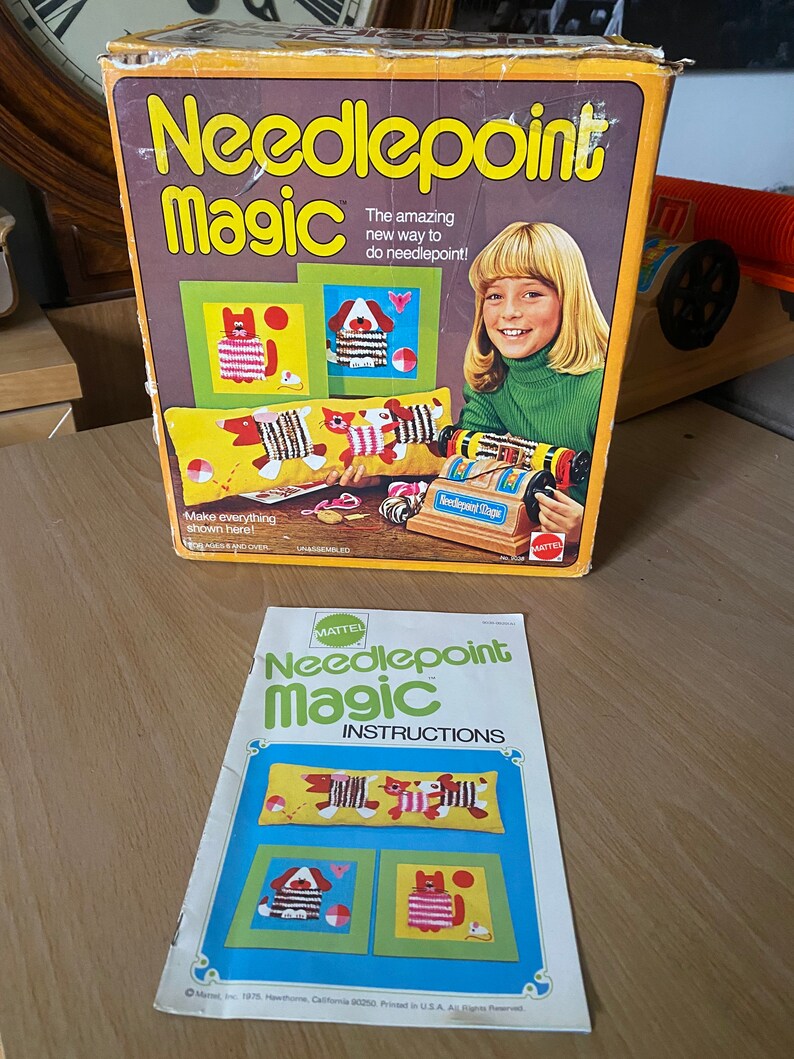 May include: A vintage Mattel Needlepoint Magic craft kit with a colorful box featuring a girl and a needlepoint loom. The box includes instructions for making needlepoint projects, including a pillow with a cat and dog design.