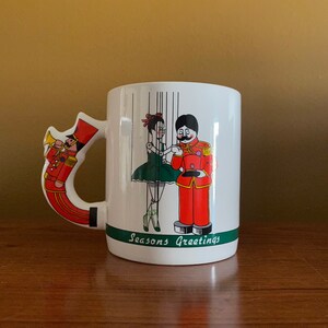 May include: White ceramic mug with a green and white handle shaped like a toy soldier playing a trumpet. The mug features a cartoon illustration of a puppet show with a female puppet in a green dress and a male puppet in a red uniform. The text "Seasons Greetings" is printed on the bottom of the mug.
