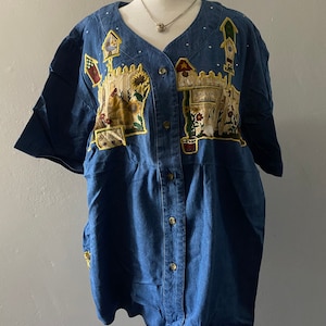 May include: A blue denim button-down shirt with short sleeves and a white polka dot pattern. The shirt features an embroidered design of a backyard garden with a fence, flowers, and a birdhouse.