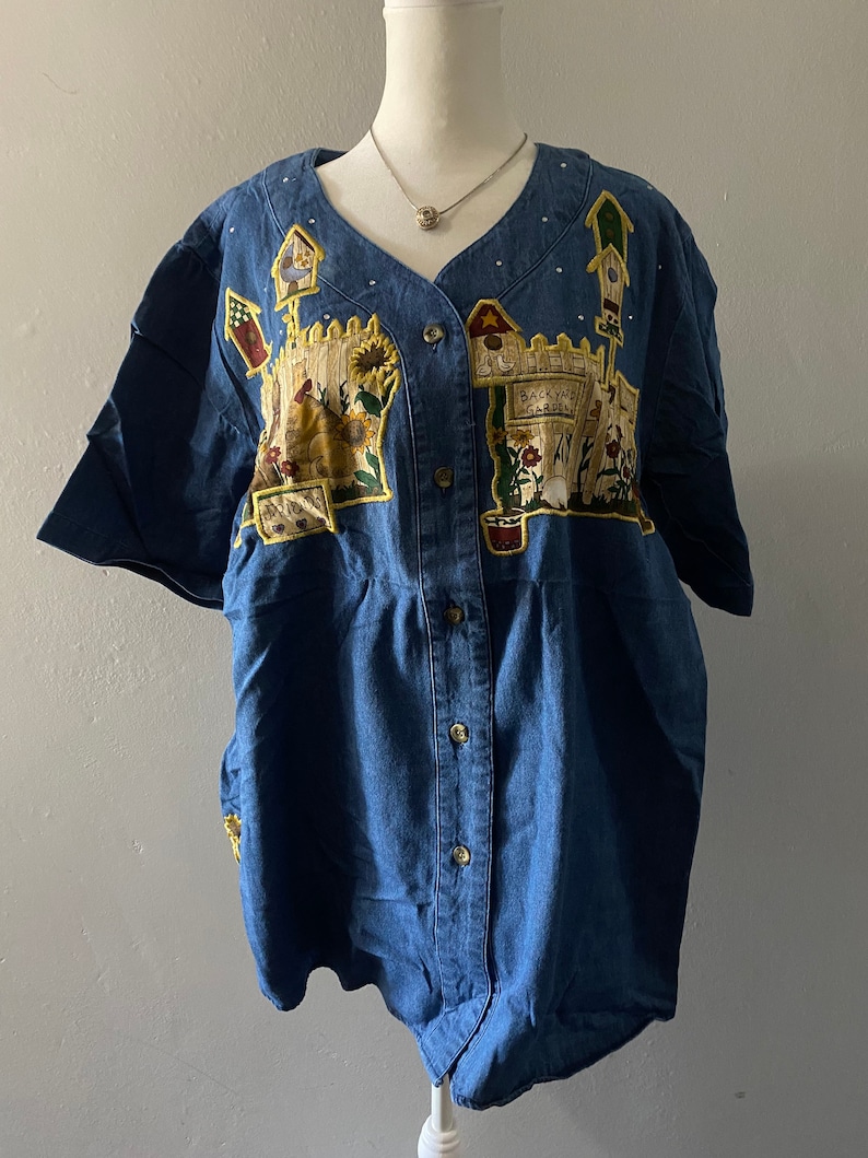 May include: A blue denim button-down shirt with short sleeves and a white polka dot pattern. The shirt features an embroidered design of a backyard garden with a fence, flowers, and a birdhouse.