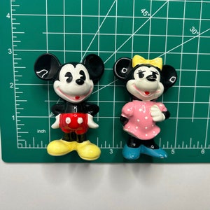 Może przedstawiać: Para ceramicznych figurek przedstawiających Myszki Miki i Minnie. Miki ma na sobie czerwone spodenki i ż&oacute;łte buty. Minnie ma na sobie r&oacute;żową sukienkę w białe kropki i niebieskie buty.