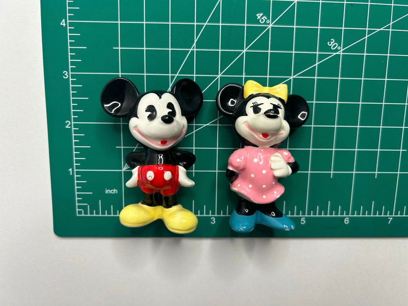 Może przedstawiać: Para ceramicznych figurek przedstawiających Myszki Miki i Minnie. Miki ma na sobie czerwone spodenki i ż&oacute;łte buty. Minnie ma na sobie r&oacute;żową sukienkę w białe kropki i niebieskie buty.