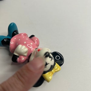 Może przedstawiać: Ceramiczna figurka Minnie Mouse. Ma na sobie r&oacute;żową sukienkę w białe kropki, niebieskie buty i ż&oacute;łtą kokardkę. Jej twarz jest pomalowana czarnymi oczami, czerwonym nosem i białym uśmiechem.