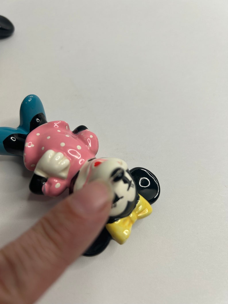 Może przedstawiać: Ceramiczna figurka Minnie Mouse. Ma na sobie r&oacute;żową sukienkę w białe kropki, niebieskie buty i ż&oacute;łtą kokardkę. Jej twarz jest pomalowana czarnymi oczami, czerwonym nosem i białym uśmiechem.