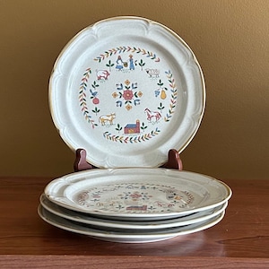 May include: Set of three white ceramic dinner plates with a farmhouse design featuring a red barn, animals, and flowers. The plates have a brown rim and a beige background.
