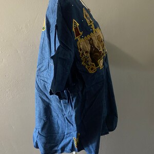 May include: A blue denim shirt with embroidered patches featuring a house, flowers, and birds. The shirt has a scoop neck and short sleeves.