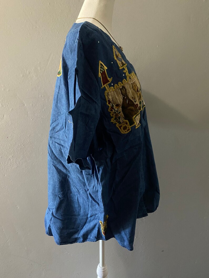 May include: A blue denim shirt with embroidered patches featuring a house, flowers, and birds. The shirt has a scoop neck and short sleeves.