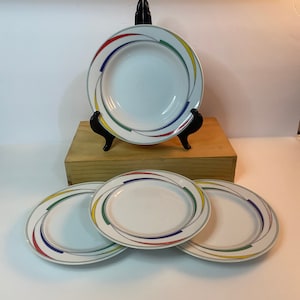 May include: Set of four white ceramic plates with a colorful abstract design. The design features red, yellow, green, and blue lines that swirl around the edge of the plates.