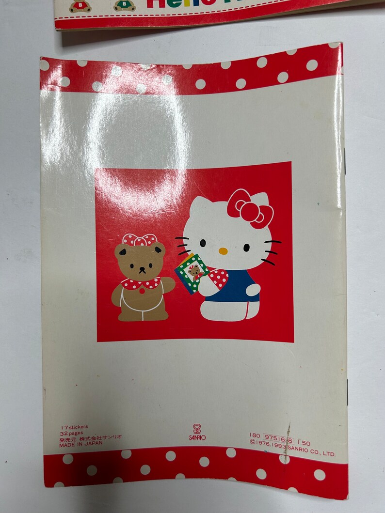 May include: A red and white book with a Hello Kitty and a bear character on the cover. The book has 32 pages and 17 stickers. The text on the back of the book reads "17 stickers, 32 pages, 発売元 株式会社サンリオ, MADE IN JAPAN, 180 97516-8 1.50, &copy;1976.1993 SANRIO CO., LTD."