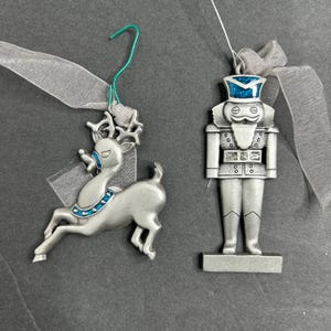 May include: Two silver Christmas ornaments: a reindeer and a nutcracker. The reindeer has blue accents and a silver ribbon. The nutcracker has a blue hat and a silver ribbon. Both ornaments are hanging against a dark gray background.