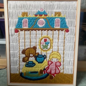 May include: A framed needlepoint picture of a teddy bear and a doll having tea in a house with a blue roof. The house is surrounded by a yellow fence and there are flowers in the window.