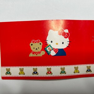 May include: A red card with a Hello Kitty illustration holding a gift box. The card has a border of small brown bears wearing different colored shirts.