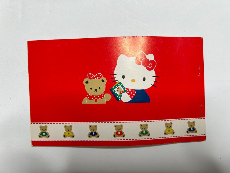 May include: A red card with a Hello Kitty illustration holding a gift box. The card has a border of small brown bears wearing different colored shirts.