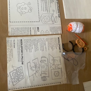 May include: A craft kit for making a needlepoint pillow. The kit includes a needlepoint canvas, yarn in various colors, a needlepoint tool, and instructions. The instructions show how to create a flower design.