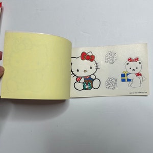 May include: A small, open book with a yellow page and a white page with a sticker of Hello Kitty holding a gift box. The sticker is on the right page. The left page is blank. The right page also has a sticker of a white bear holding a gift box. The text on the sticker reads "&copy;'76,'81 SANRIO CO., LTD."