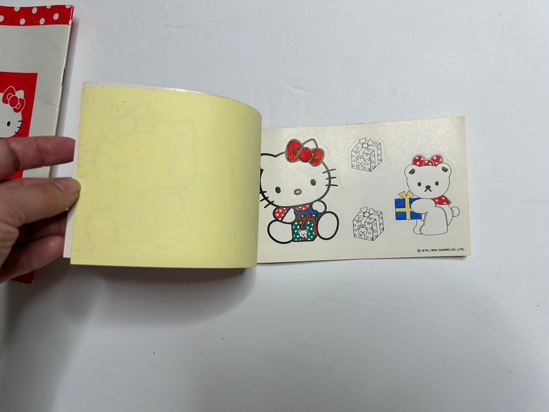 May include: A small, open book with a yellow page and a white page with a sticker of Hello Kitty holding a gift box. The sticker is on the right page. The left page is blank. The right page also has a sticker of a white bear holding a gift box. The text on the sticker reads "&copy;'76,'81 SANRIO CO., LTD."