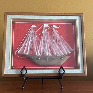 May include: A framed string art piece depicting a sailboat with white string and a wooden base. The frame is brown wood with a light blue mat and a red background.