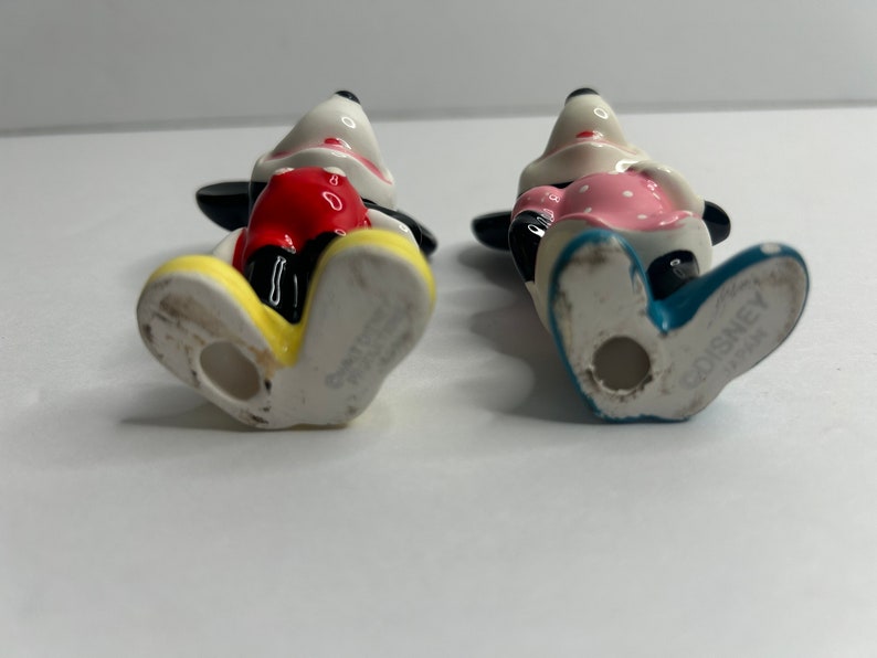 Może przedstawiać: Para ceramicznych figurek Mickey Mouse'a i Minnie Mouse. Mickey ma na sobie czerwone spodenki i ż&oacute;łte buty. Minnie ma na sobie r&oacute;żową sukienkę w białe kropki i niebieskie buty. Obie figurki mają napis "Disney Japan" na spodzie st&oacute;p.
