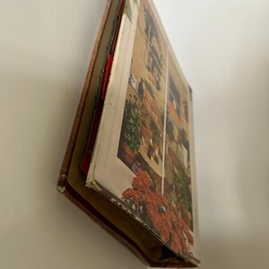 May include: A vintage book with a colorful illustration of a window scene with potted plants and flowers. The book's cover and pages are visible, showcasing a detailed, aged design. The book is leaning against a white wall.