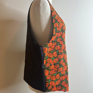 May include: A black sleeveless apron with a pumpkin print on the right side. The apron has a black fabric on the left side and a pumpkin print on the right side. The pumpkin print is orange and green and features a pattern of jack-o'-lantern faces.