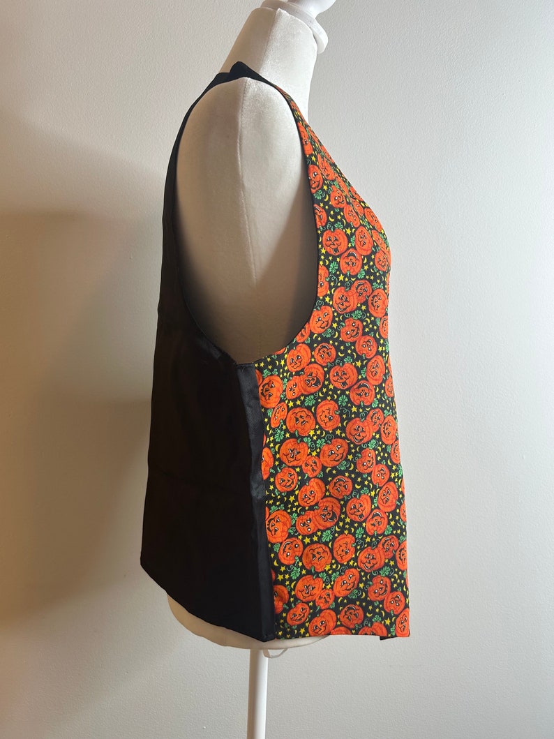 May include: A black sleeveless apron with a pumpkin print on the right side. The apron has a black fabric on the left side and a pumpkin print on the right side. The pumpkin print is orange and green and features a pattern of jack-o'-lantern faces.