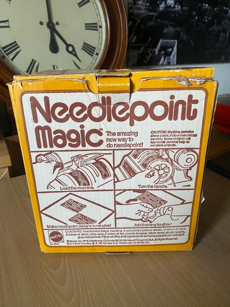 May include: A yellow box with the words "Needlepoint Magic" in brown letters. The box shows a toy machine that makes needlepoint designs. The text on the box says "The amazing new way to do needlepoint!" and "Make needlepoint designs in minutes!"  The box is from Mattel.