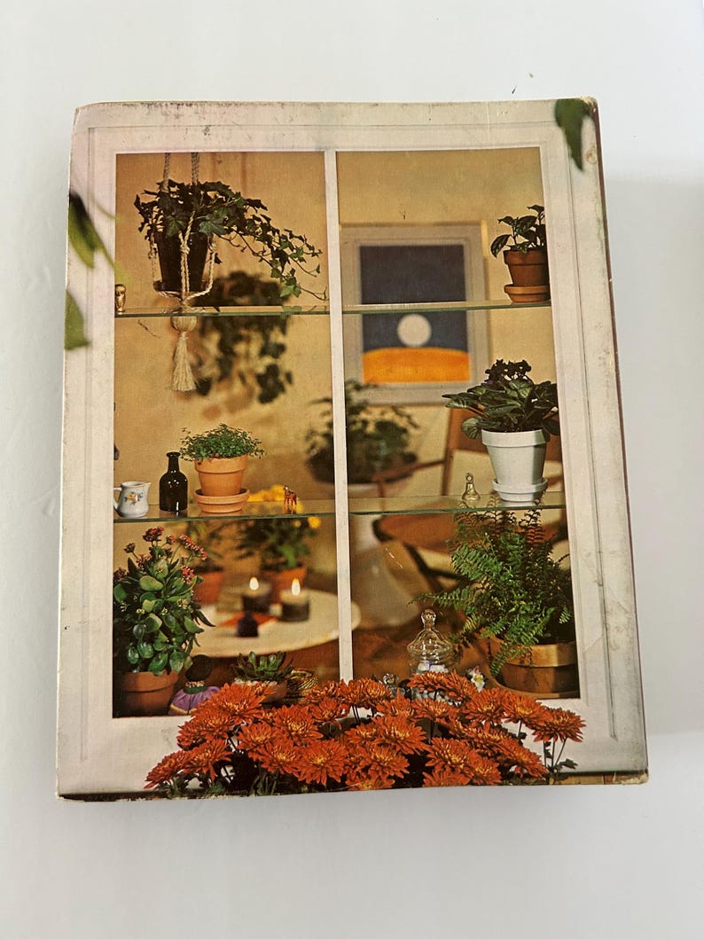 May include: A vintage book cover featuring a window display with potted plants on glass shelves. The scene includes hanging plants, a painting, and orange chrysanthemums. The overall aesthetic is warm and inviting, with a focus on indoor gardening.