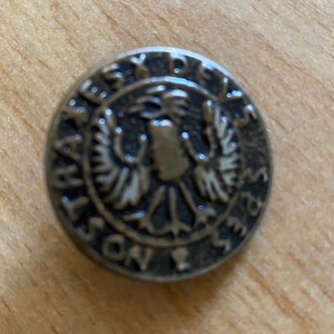 May include: A round, bronze-colored button with a raised design of a bird with outstretched wings. The button has the text "REX DELS" around the edge.