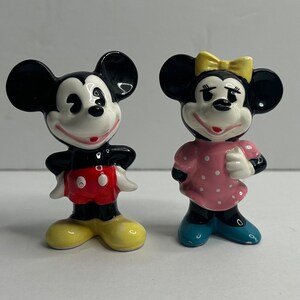 Może przedstawiać: Para ceramicznych figurek przedstawiających Myszki Miki i Minnie. Miki ma na sobie swoje charakterystyczne czerwone spodenki i ż&oacute;łte buty. Minnie ma na sobie r&oacute;żową sukienkę w kropki i niebieskie buty.