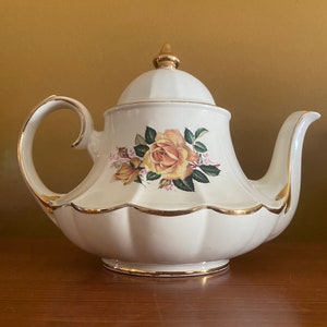 Op de afbeelding: Een witte theepot met een gouden rand en een bloemenontwerp met gele rozen. De theepot heeft een gegolfde basis en een deksel met een gouden knop.