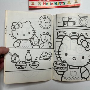 May include: A black and white coloring book featuring Hello Kitty. The cover shows Hello Kitty baking a cake with a friend. The book includes a clock, a scale, and other kitchen items.
