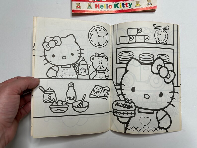May include: A black and white coloring book featuring Hello Kitty. The cover shows Hello Kitty baking a cake with a friend. The book includes a clock, a scale, and other kitchen items.