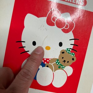 May include: A red and white Hello Kitty coloring book with stickers. The cover features Hello Kitty holding a brown teddy bear. The book has the words "Hello Kitty" printed on the bottom in colorful letters.