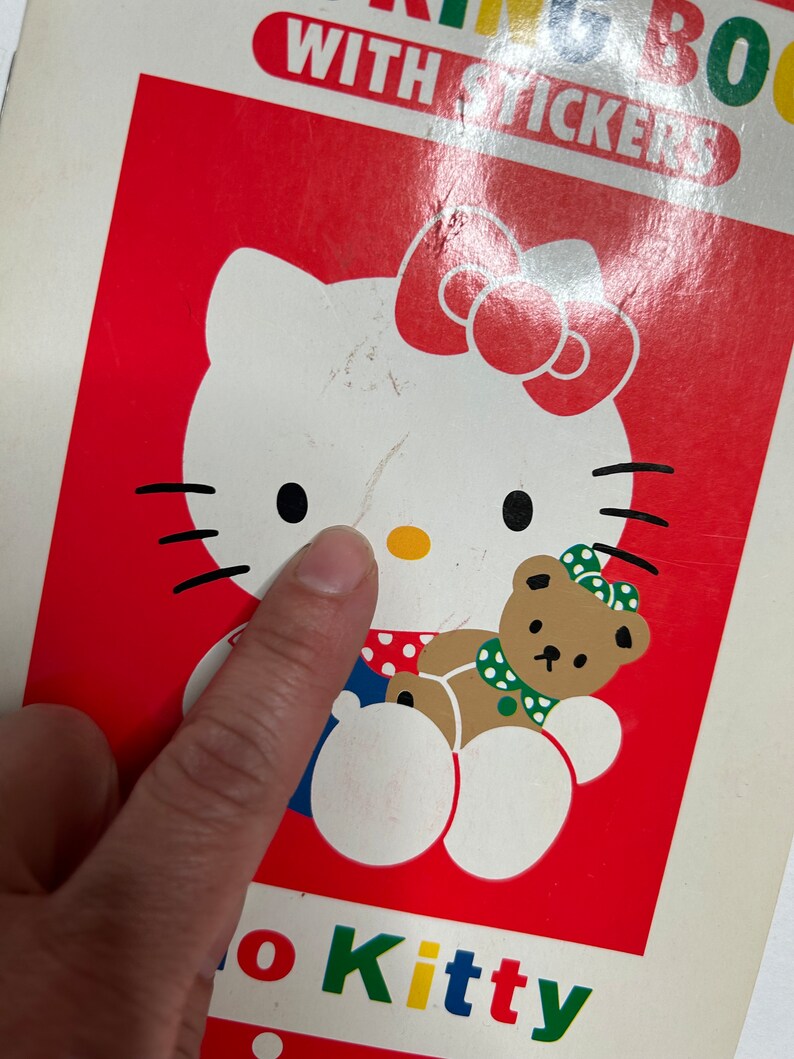 May include: A red and white Hello Kitty coloring book with stickers. The cover features Hello Kitty holding a brown teddy bear. The book has the words "Hello Kitty" printed on the bottom in colorful letters.