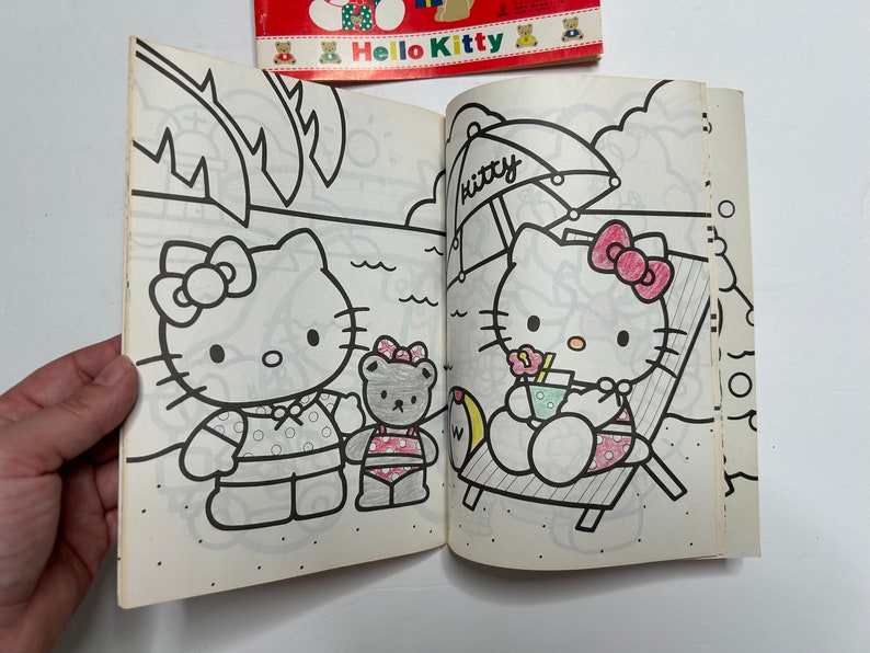 May include: A coloring book featuring Hello Kitty on the cover. The inside pages show two black and white illustrations of Hello Kitty, one with a beach umbrella and the other with a friend.