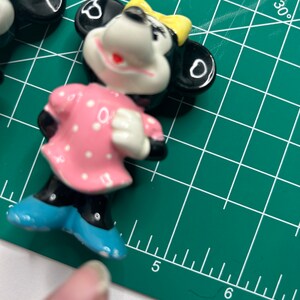 Może przedstawiać: Ceramiczną figurkę przedstawiającą mysz z kresk&oacute;wek w r&oacute;żowej sukience w białe kropki, niebieskich butach, ż&oacute;łtej kokardce i czarnych uszach.