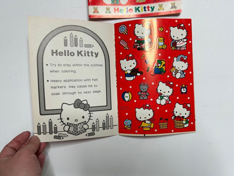May include: A coloring book featuring Hello Kitty. The cover is red with white polka dots and features Hello Kitty in various poses. The inside of the book has a black and white illustration of Hello Kitty and instructions for coloring. The text reads "Try to stay within the outlines when coloring. Heavy application with felt markers may cause ink to soak through to next page."