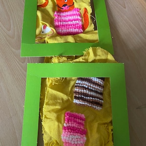 May include: Two green picture frames with yellow fabric inside. The fabric has colorful yarn pom-poms attached to it. One frame has a cat with a pink and white striped sweater. The other frame has a pink and white pom-pom and a brown and white striped pom-pom.