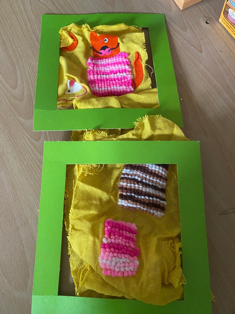 May include: Two green picture frames with yellow fabric inside. The fabric has colorful yarn pom-poms attached to it. One frame has a cat with a pink and white striped sweater. The other frame has a pink and white pom-pom and a brown and white striped pom-pom.